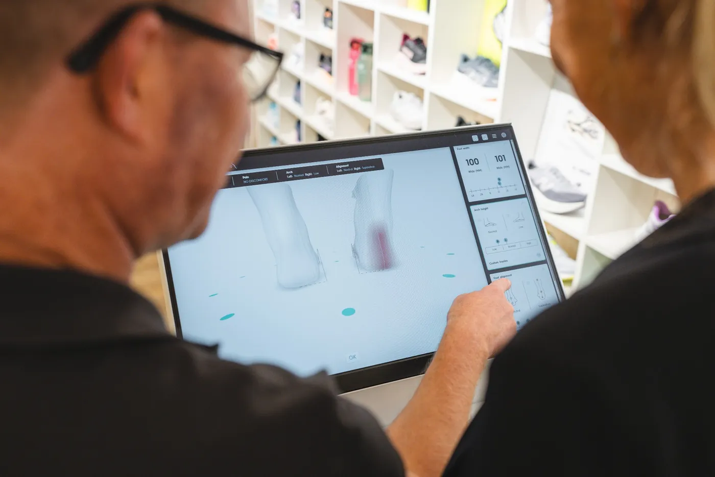 Een medewerker wijst op het scherm van de 3D-voetscan de stand van de voeten aan, naast de voetbreedte en de hoogte van de voetboog